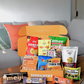 An open MUNCHEES orange snack box filled with assorted packaged snacks sits on a wooden table in a living room setting. Visible brands and snack packages include Meiji milk chocolate, Calbee Ultra Spicy Potato Chips, and others.