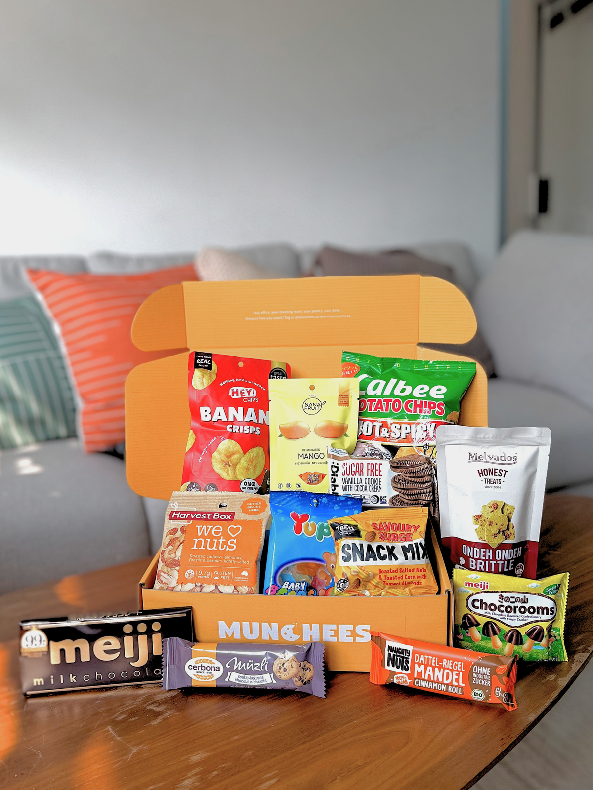 An open MUNCHEES orange snack box filled with assorted packaged snacks sits on a wooden table in a living room setting. Visible brands and snack packages include Meiji milk chocolate, Calbee Ultra Spicy Potato Chips, and others.