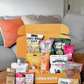 An open orange MUNCHEES snack box filled with assorted packaged snacks and placed on a wooden table in front of a living room sofa background. Visible snack brands include Hey Chips!, Marou, Zenko, Amazin' Graze, and others.