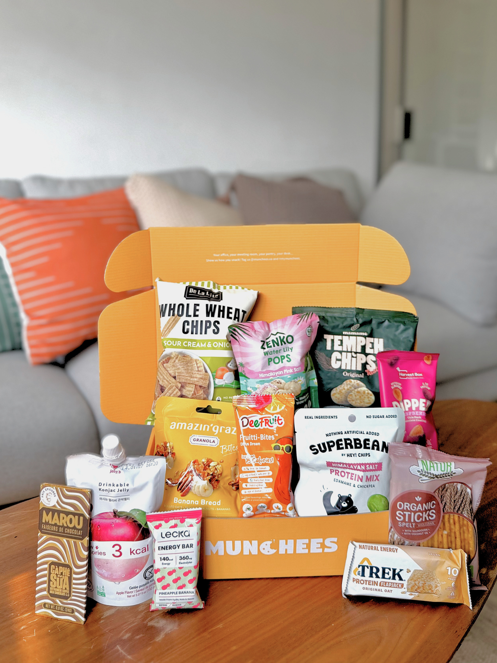 An open orange MUNCHEES snack box filled with assorted packaged snacks and placed on a wooden table in front of a living room sofa background. Visible snack brands include Hey Chips!, Marou, Zenko, Amazin' Graze, and others.