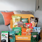 A MUNCHEES snack box filled with various packaged snacks, displayed on a wooden table in front of a couch with colorful pillows. The savoury box snack  for office pantry snack and corporate gifting