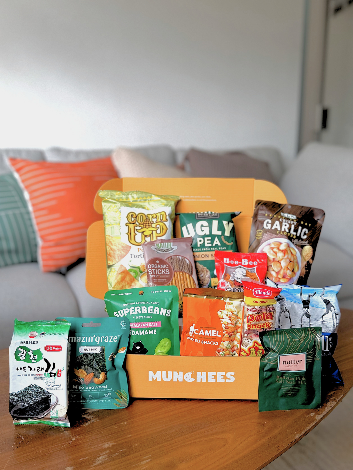 A MUNCHEES snack box filled with various packaged snacks, displayed on a wooden table in front of a couch with colorful pillows. The savoury box snack  for office pantry snack and corporate gifting
