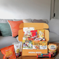 An open MUNCHEES snack box filled with assorted packaged snacks, placed on a wooden coffee table in front of a gray sofa with colorful pillows. Visible snack brands include M&M's, Kinder Bueno's, KitKat, and Zenko. The sweet box snack for office pantry snack and corporate gifting