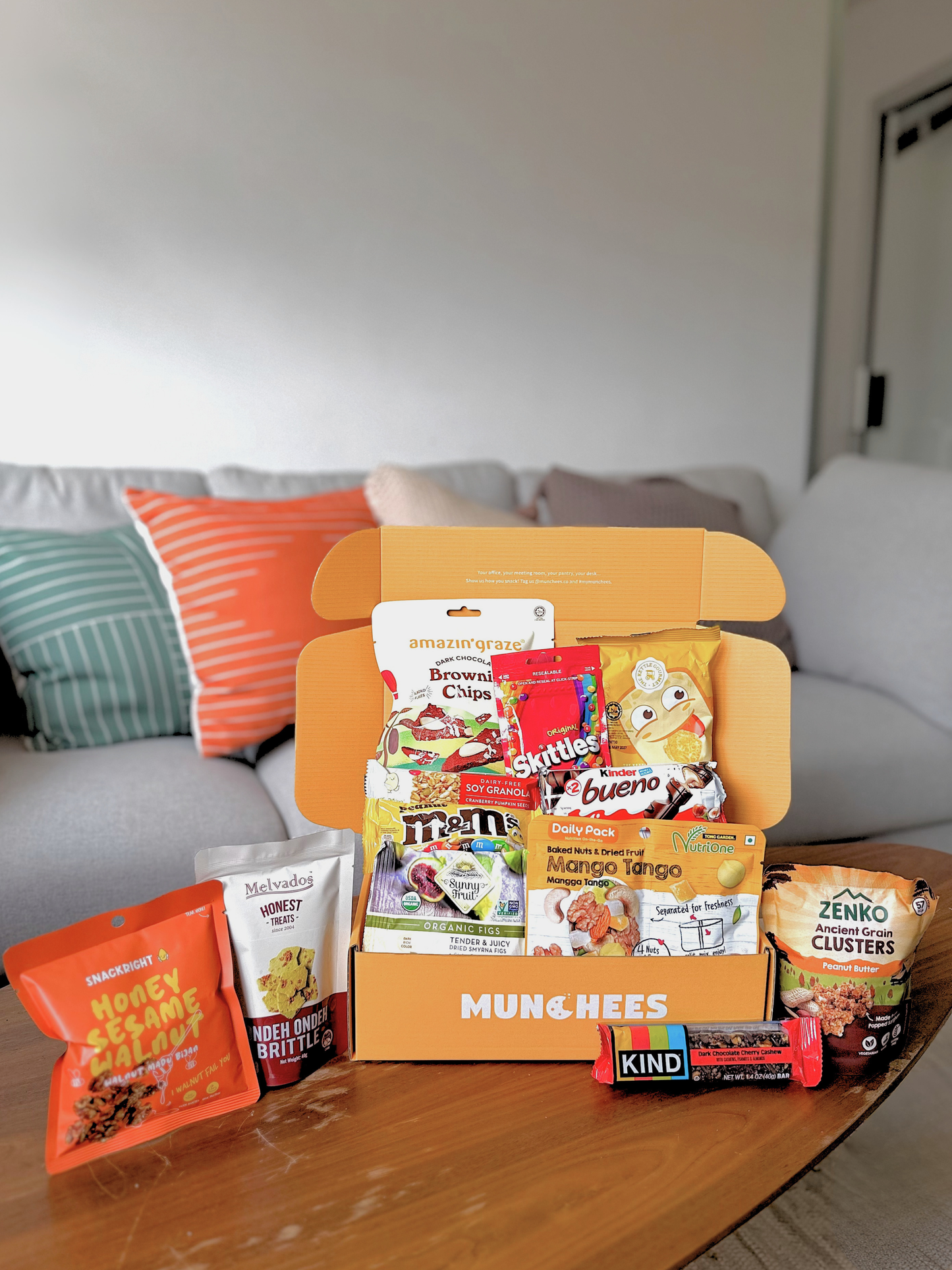 An open MUNCHEES snack box filled with assorted packaged snacks, placed on a wooden coffee table in front of a gray sofa with colorful pillows. Visible snack brands include M&M's, Kinder Bueno's, KitKat, and Zenko. The sweet box snack for office pantry snack and corporate gifting